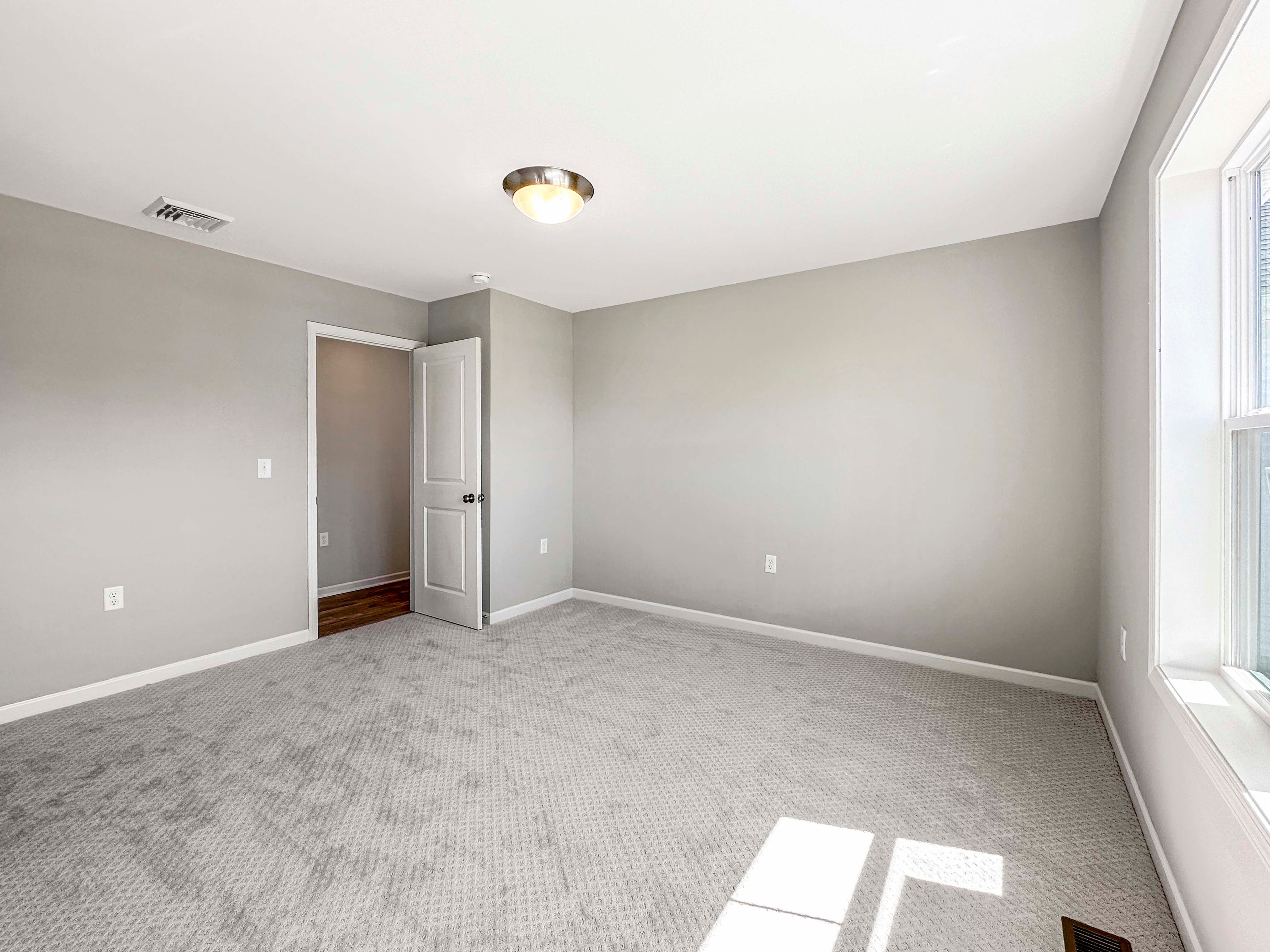 Spacious bedroom with large windows providing natural light, gray carpet, and neutral walls.