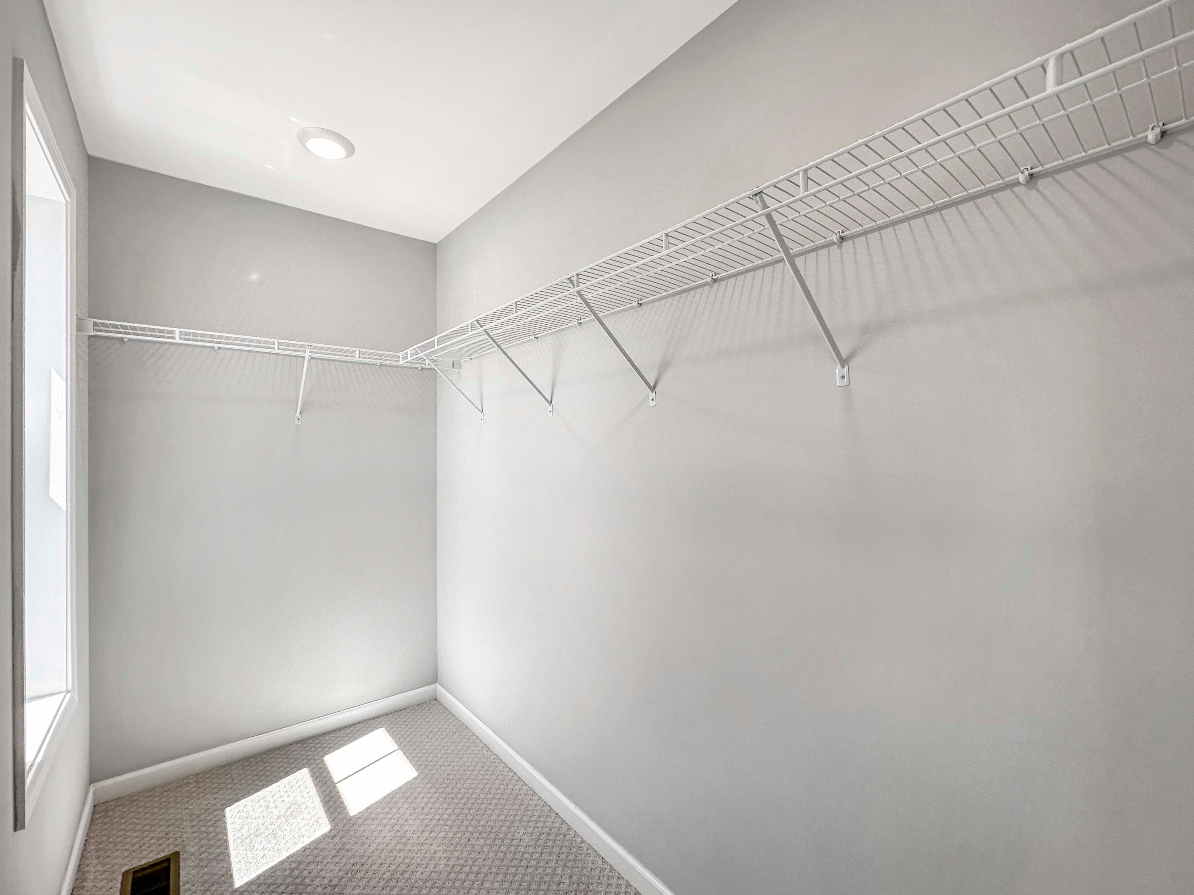 Spacious walk-in closet with wire shelving, neutral walls, and carpet flooring. Bright light from an overhead fixture.