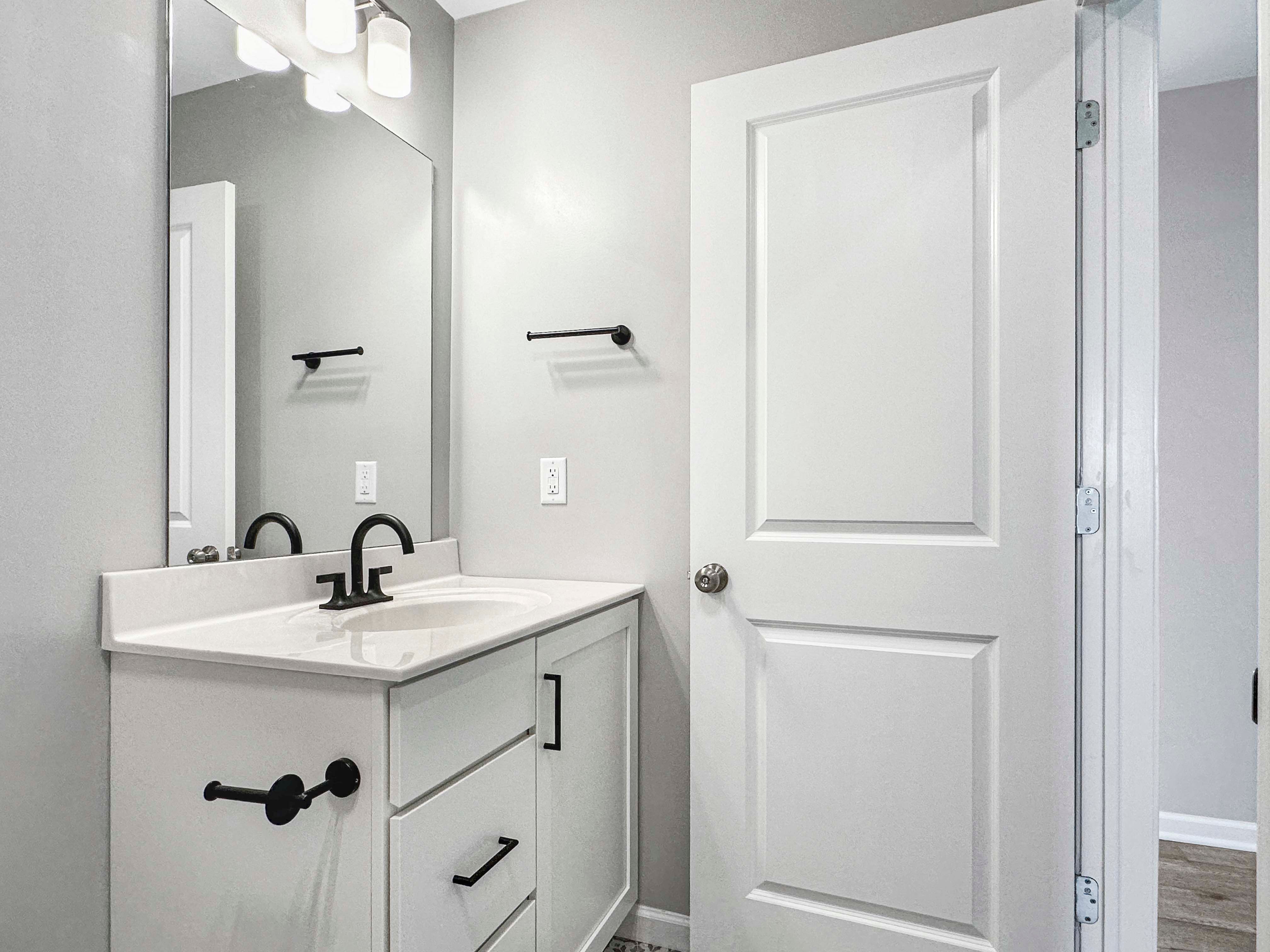 Modern bathroom with white vanity, black fixtures, and a large mirror. Simple, clean design with white door and neutral tones.