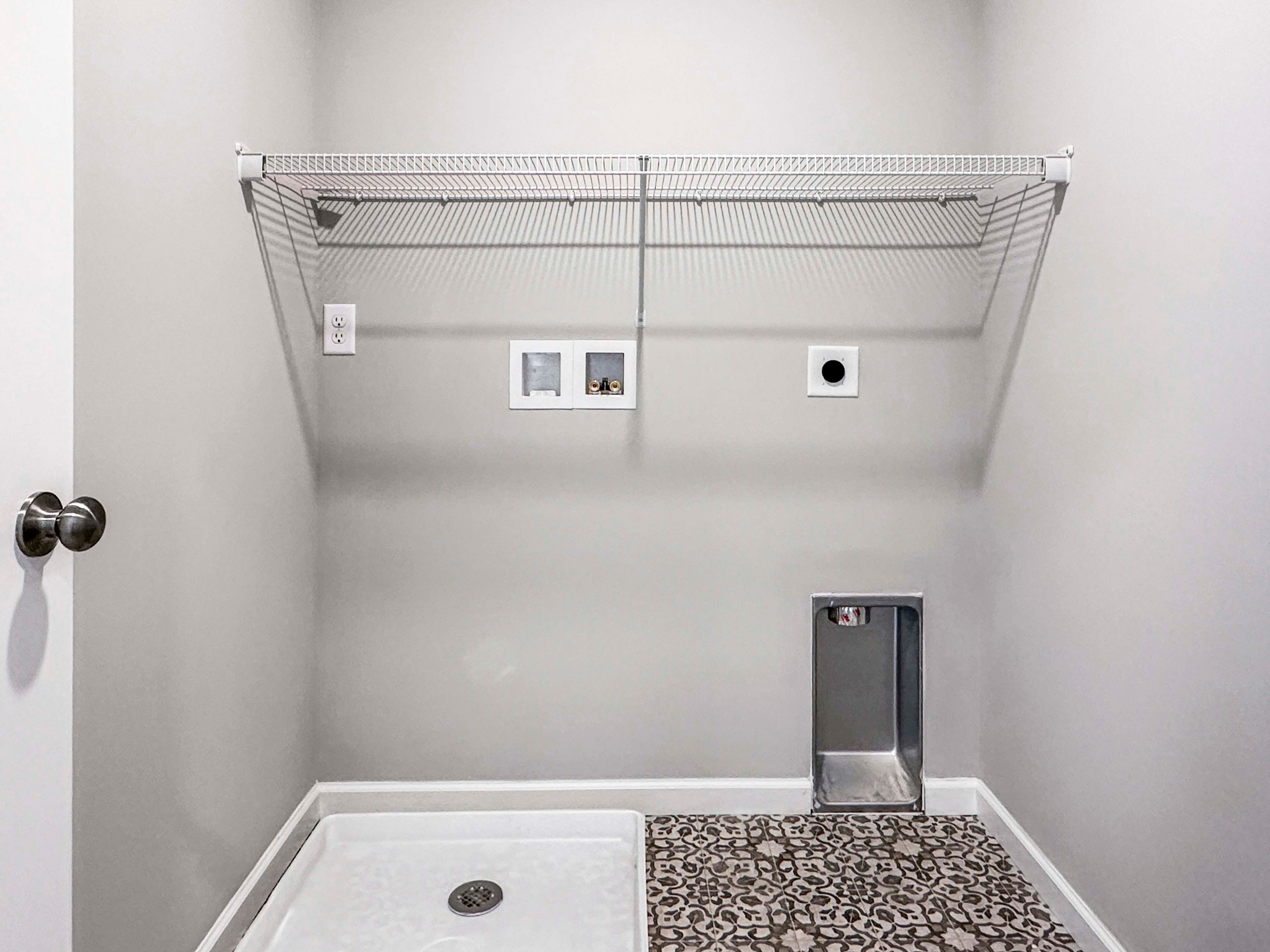Close-up of laundry room plumbing hookups, wire shelf, and patterned tile flooring, providing space for appliances.