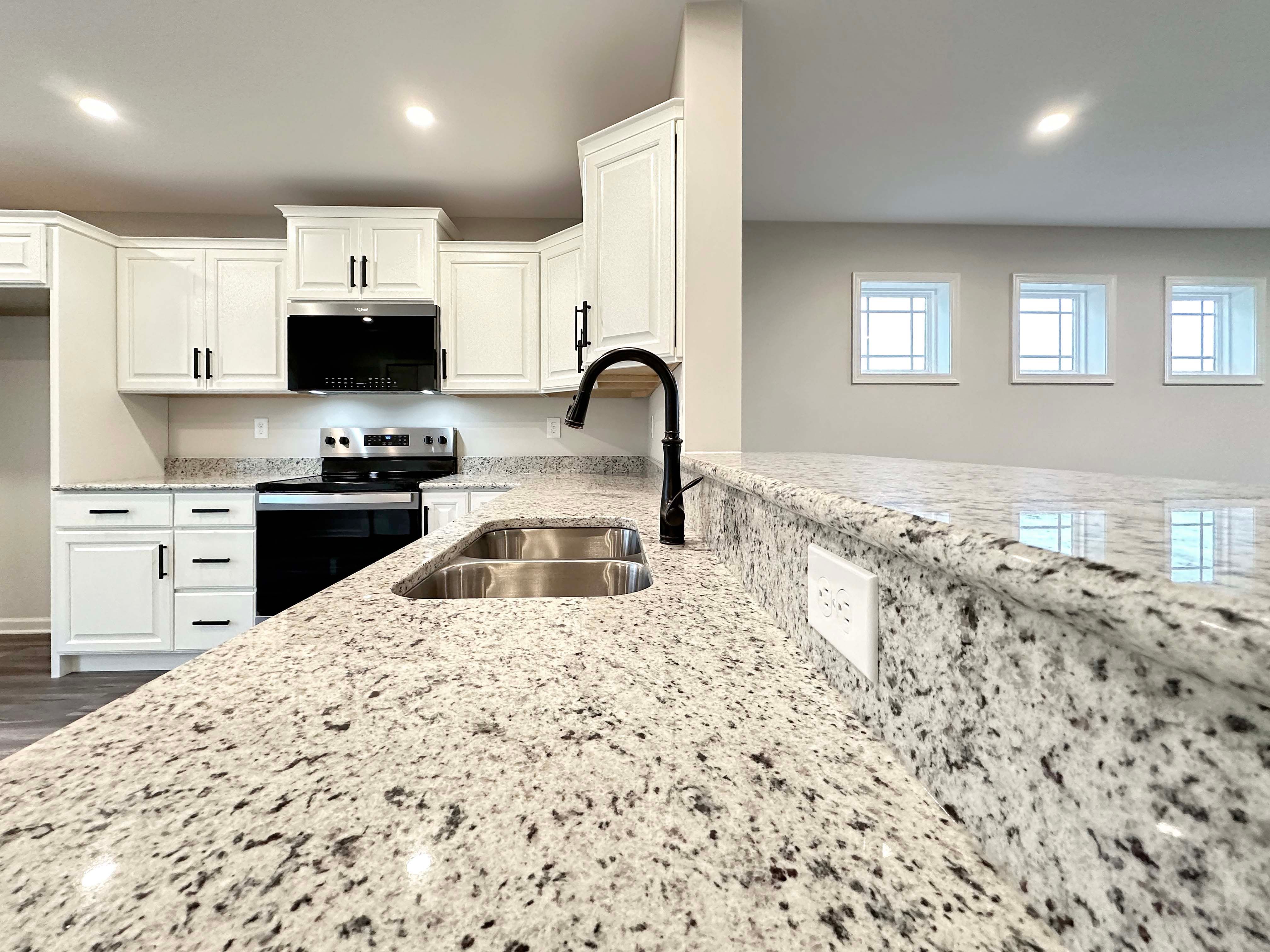 Spacious kitchen with white cabinetry, granite countertops, and stainless-steel sink. View extends into the unfurnished living room.