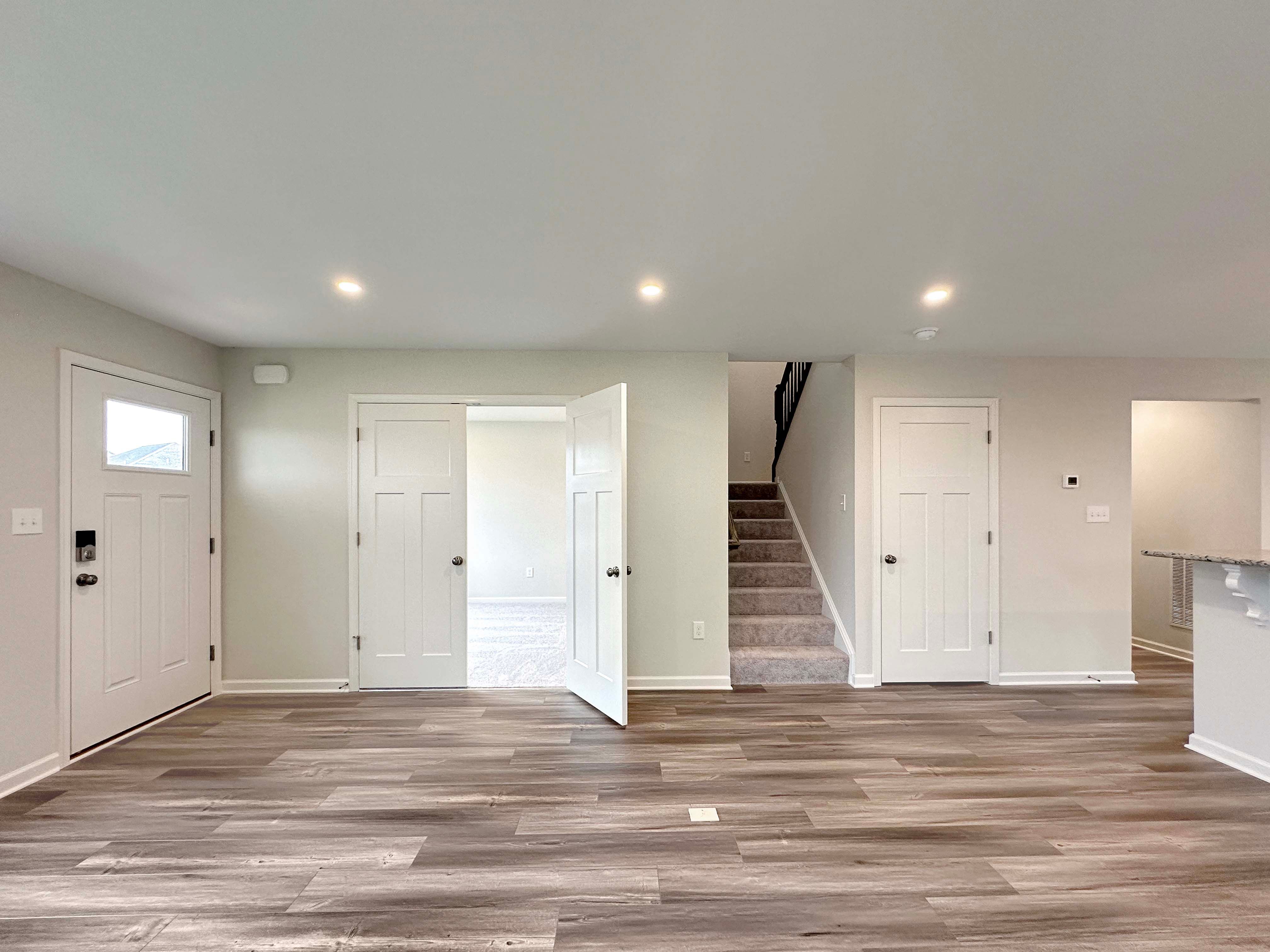 Front entry view with large windows, wood-look floors, and staircase leading upstairs, in an unfurnished living room.