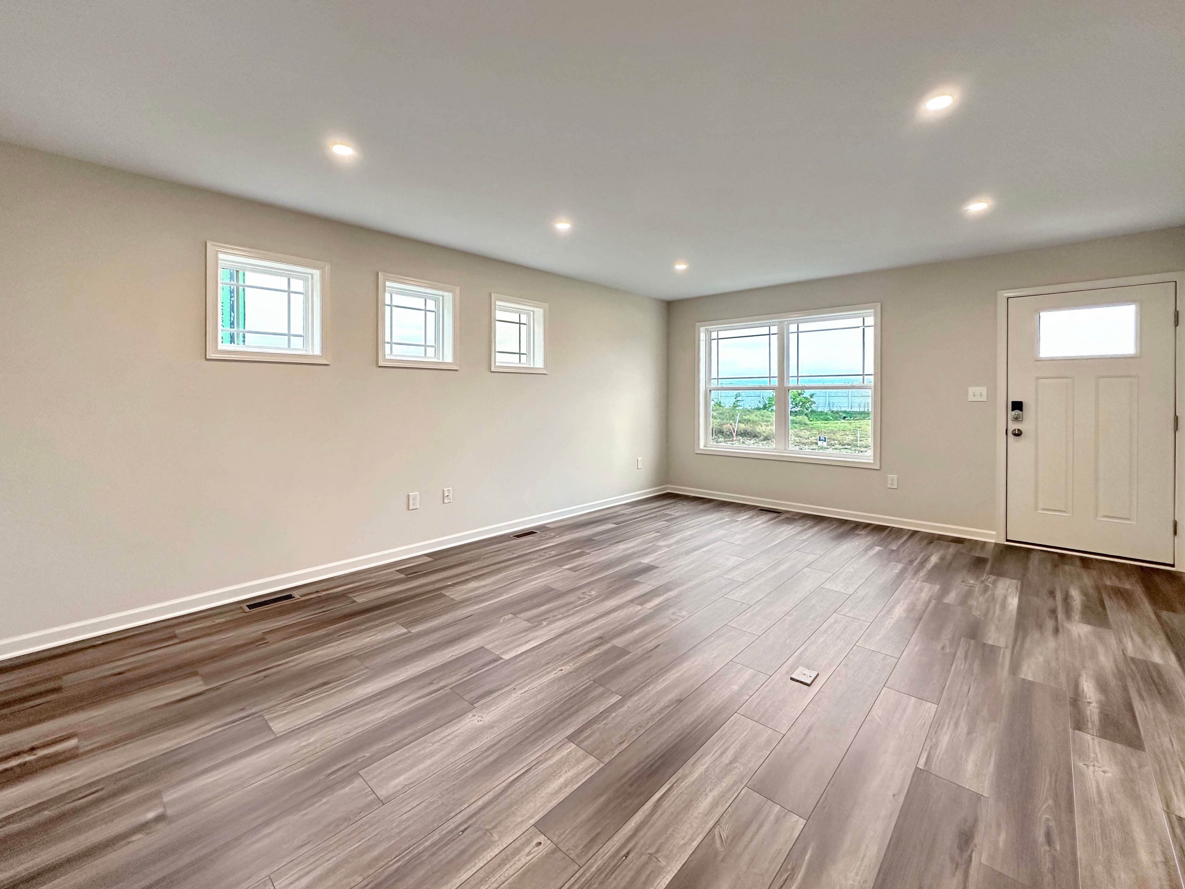 Spacious living room with wood-look flooring, large windows, and light flooding the unfurnished room.