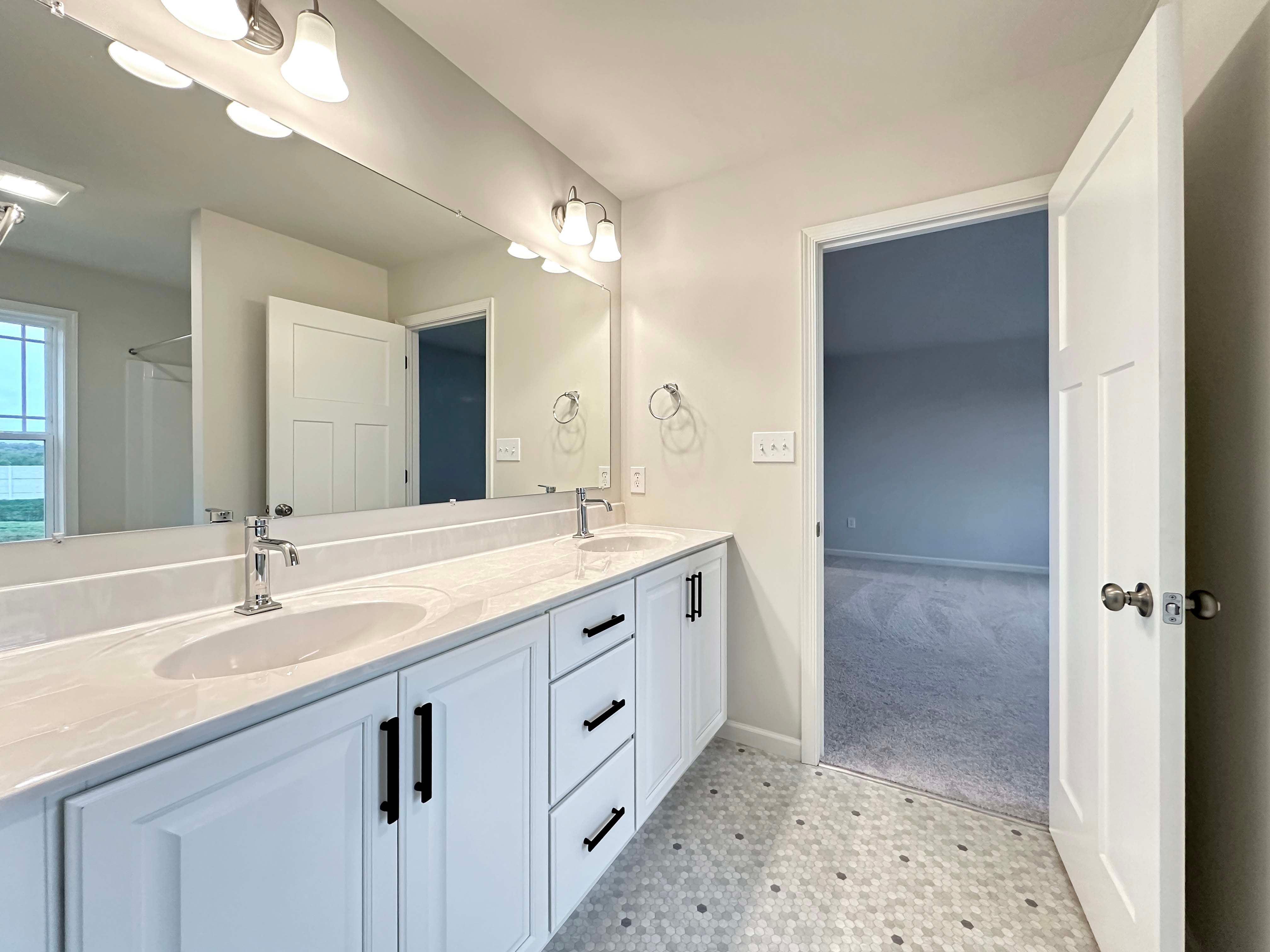 Bright bathroom with a dual-sink vanity, large mirror, and hexagon-tiled floor, leading to the bedroom