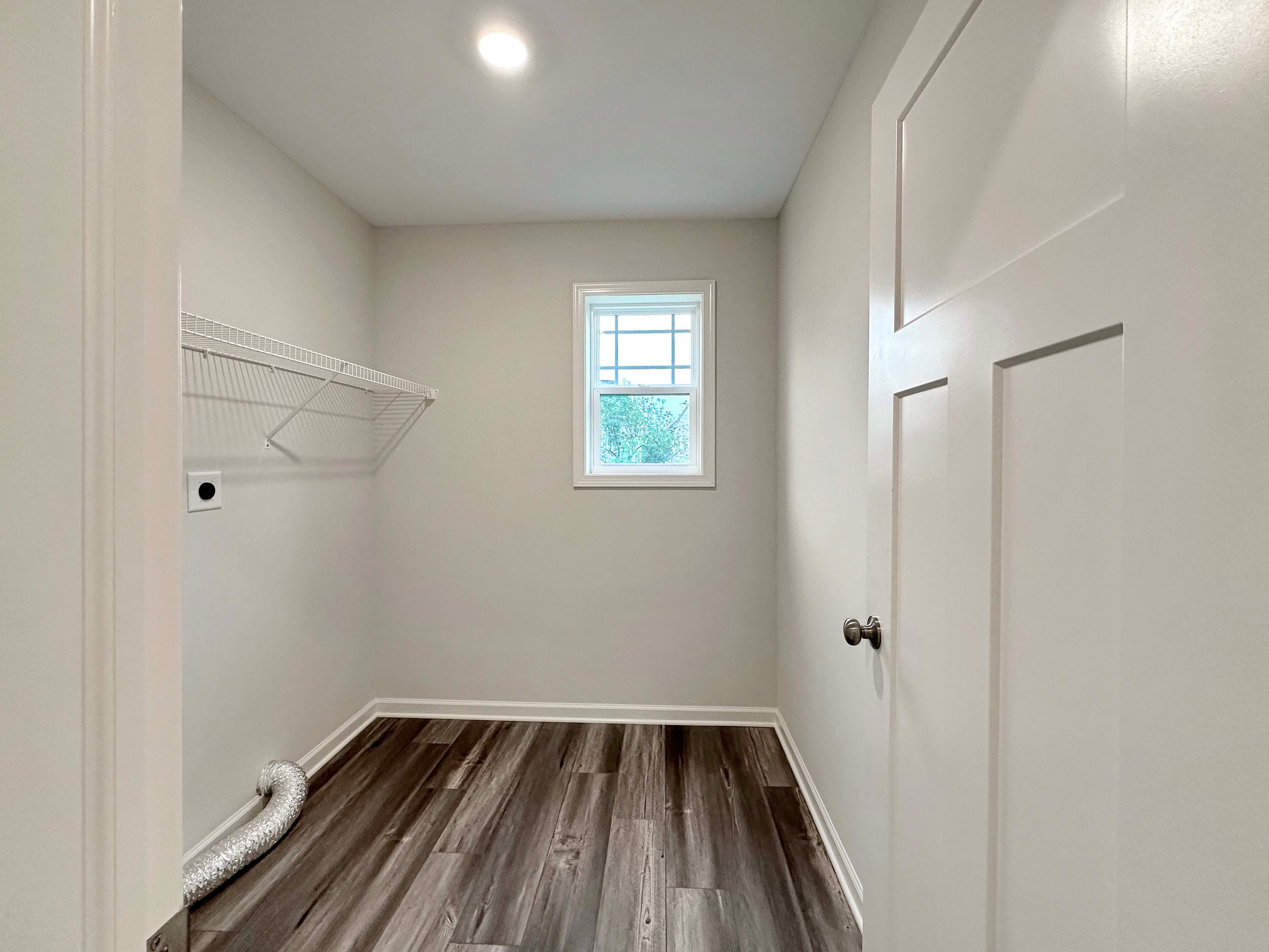 Bright laundry room with hardwood flooring, a window, and white wire shelving, ready for washer and dryer installation.