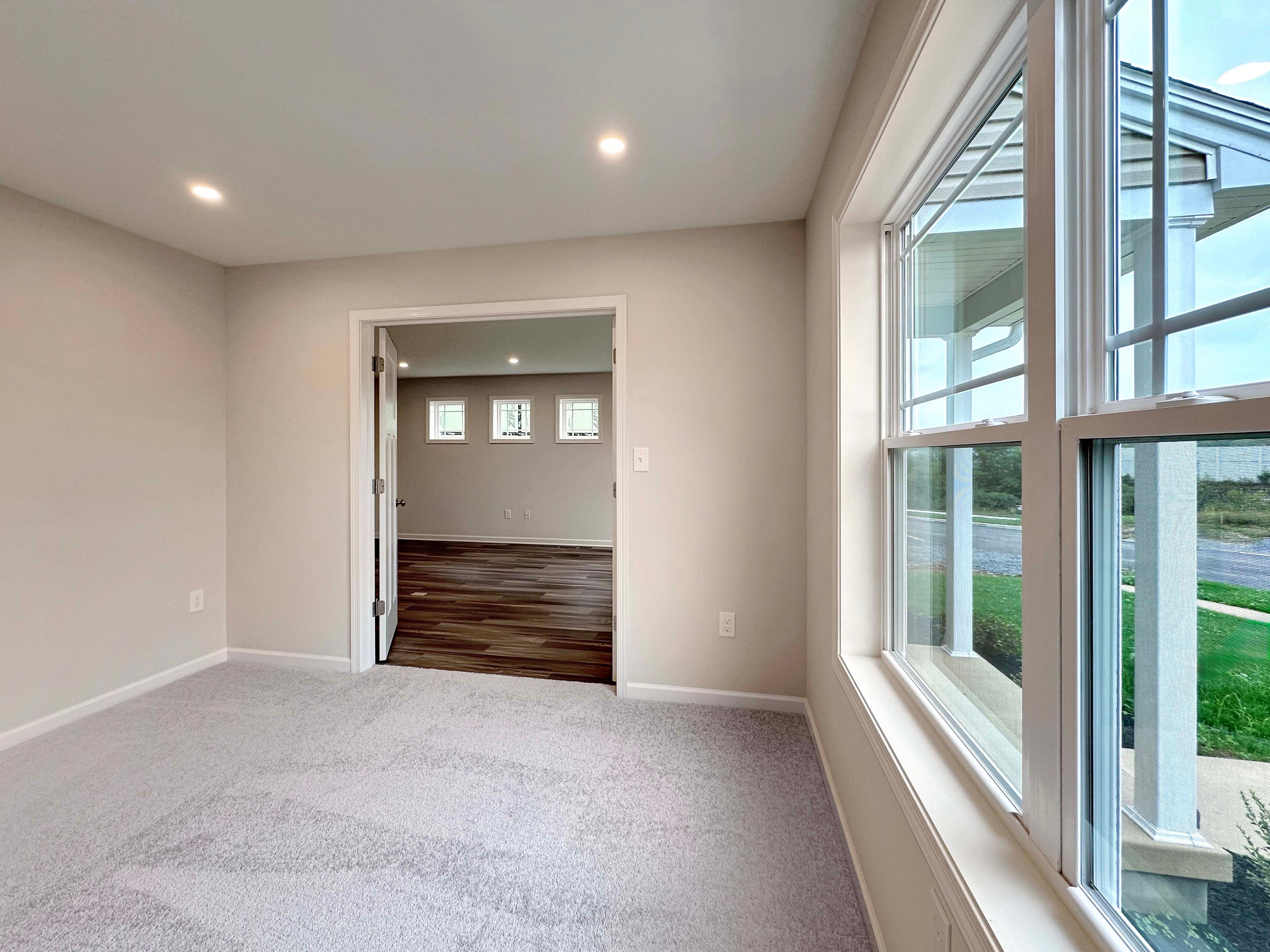 Light-filled room with large windows, carpet flooring, and view of both the front yard and side room with hardwood flooring.