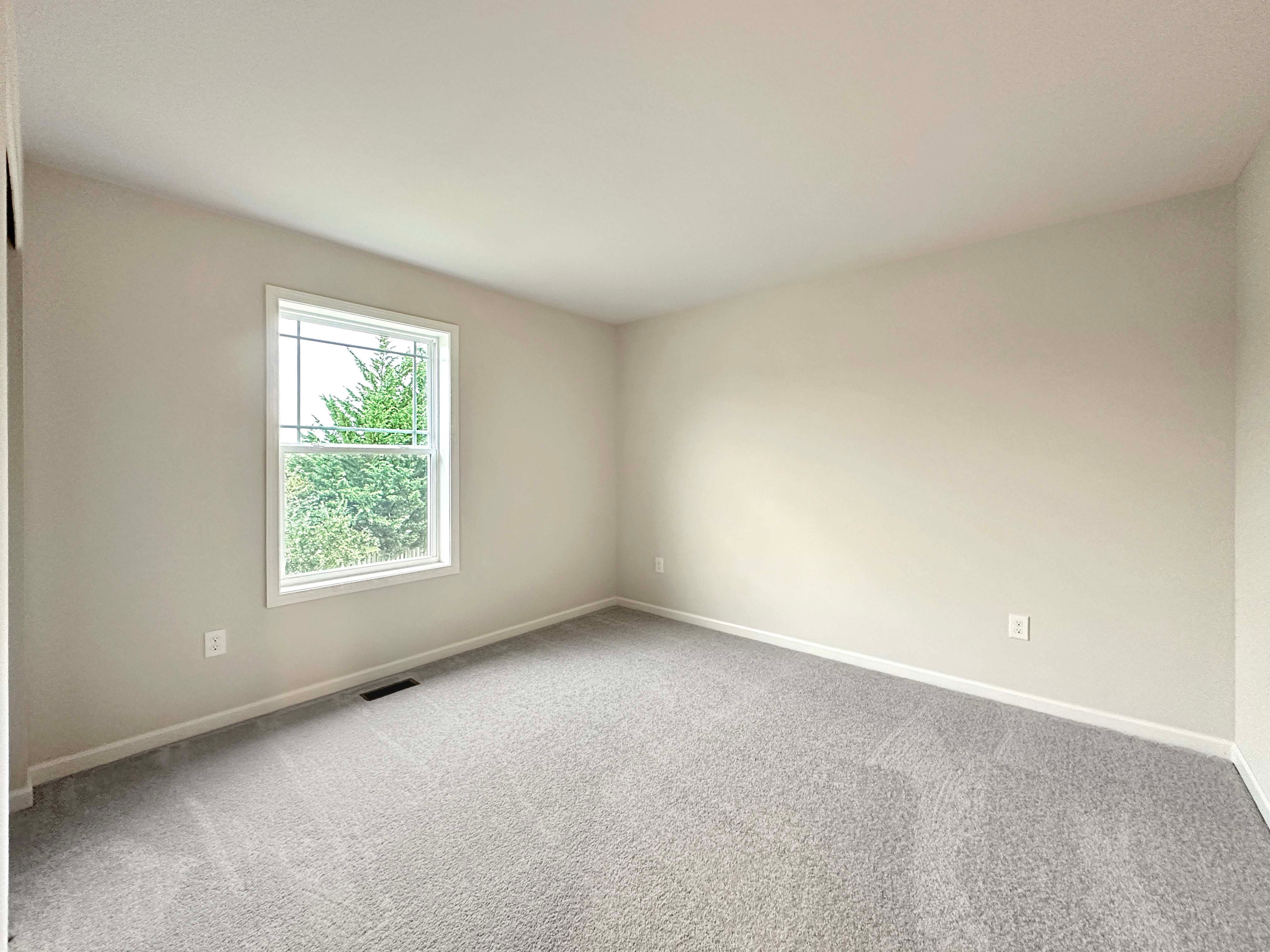 Bright bedroom with a large window, gray carpet, and neutral walls, providing a serene and open space.