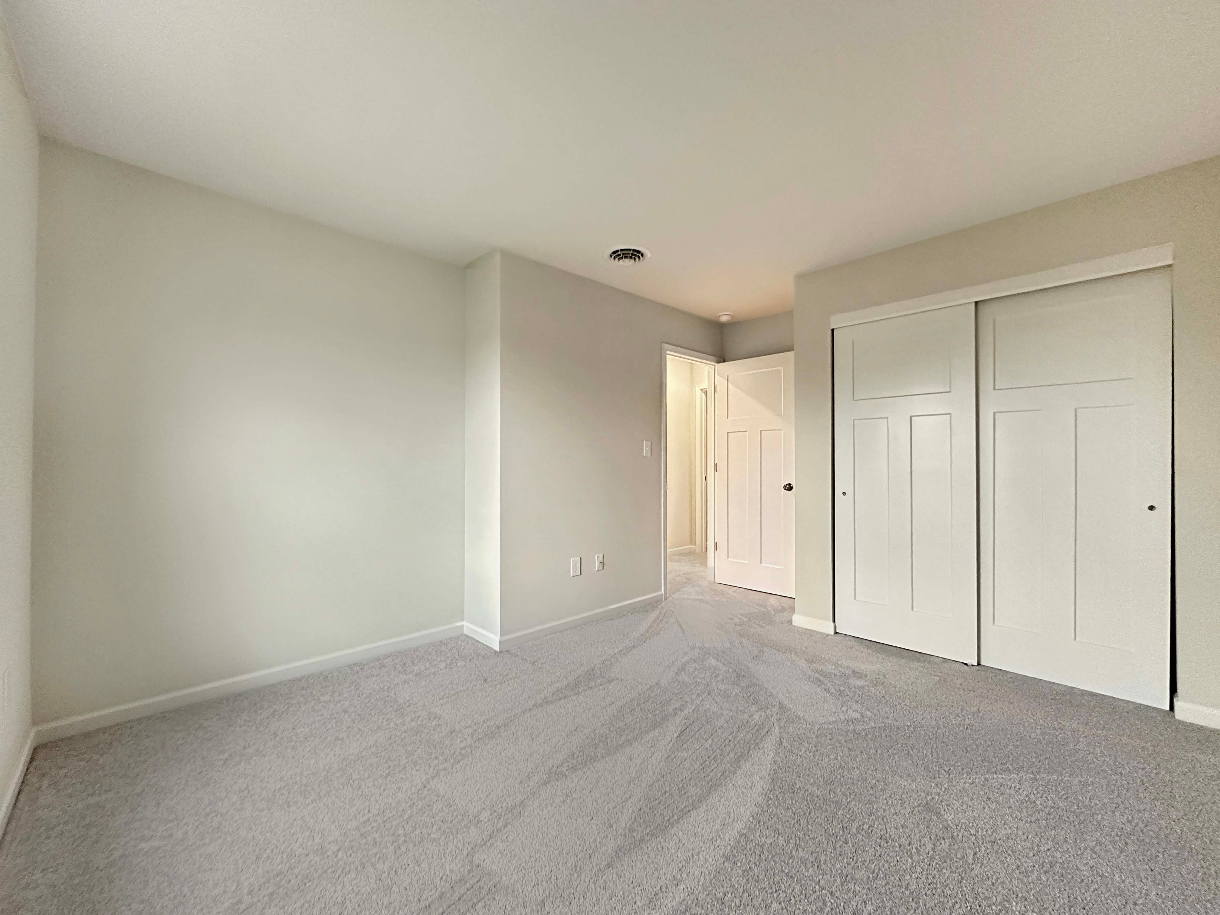 Spacious unfurnished bedroom with light gray walls, carpet flooring, and double sliding closet doors.