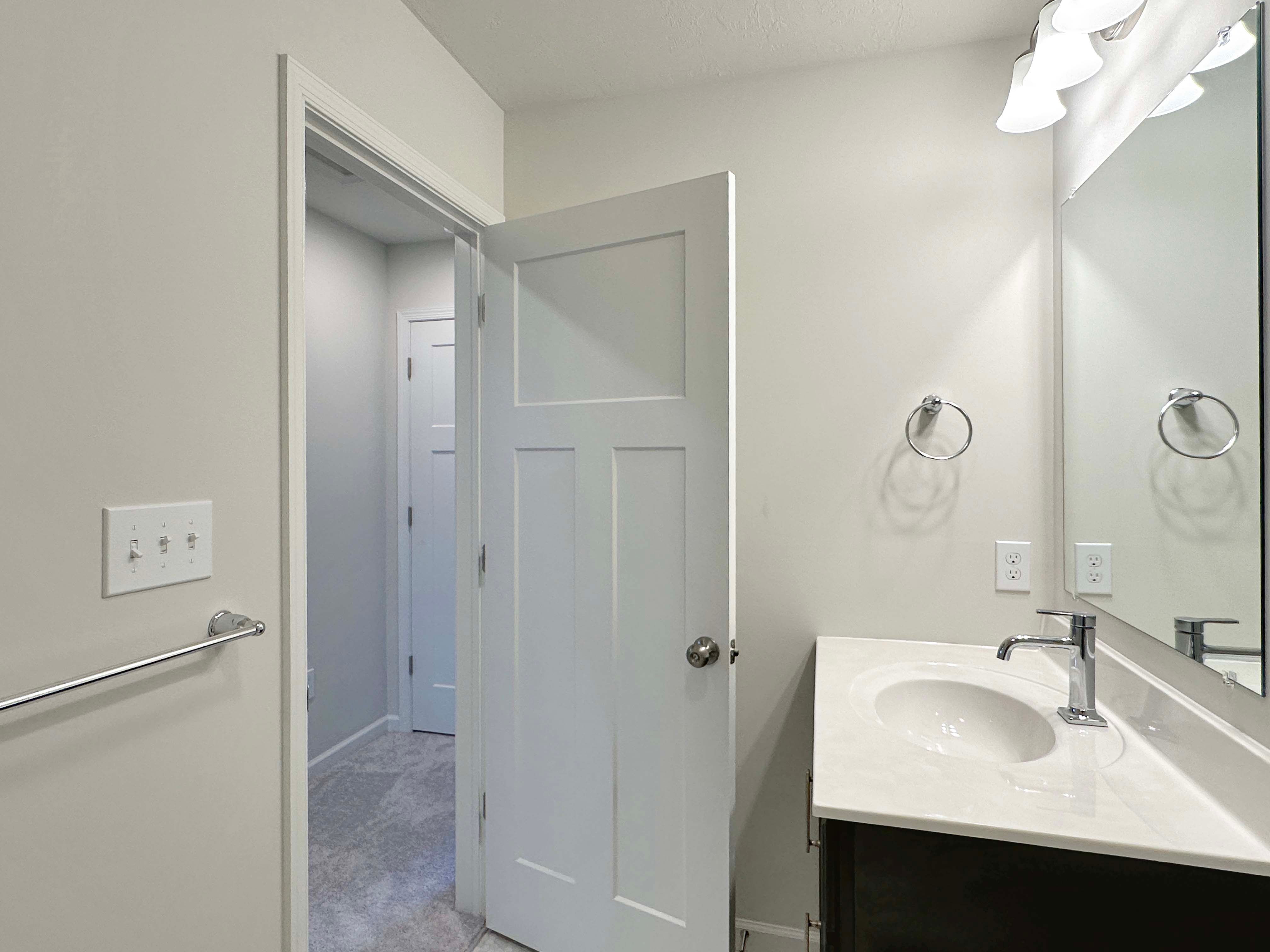Bathroom with white walls, a pedestal sink, and a mirror reflecting the hallway, leading to a carpeted area