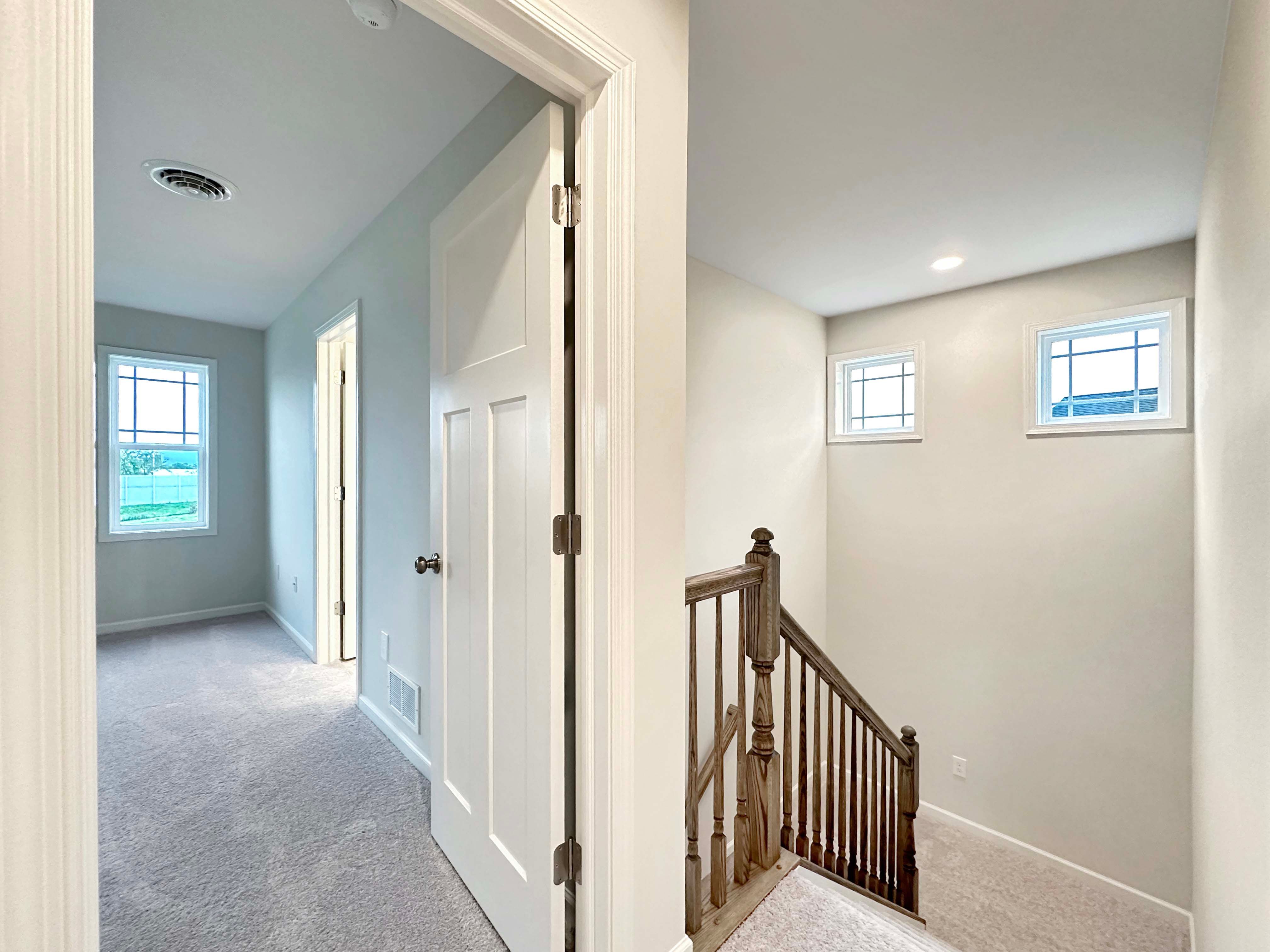 Bright upstairs hallway with two windows, carpeted flooring, and a wooden handrail leading downstairs.