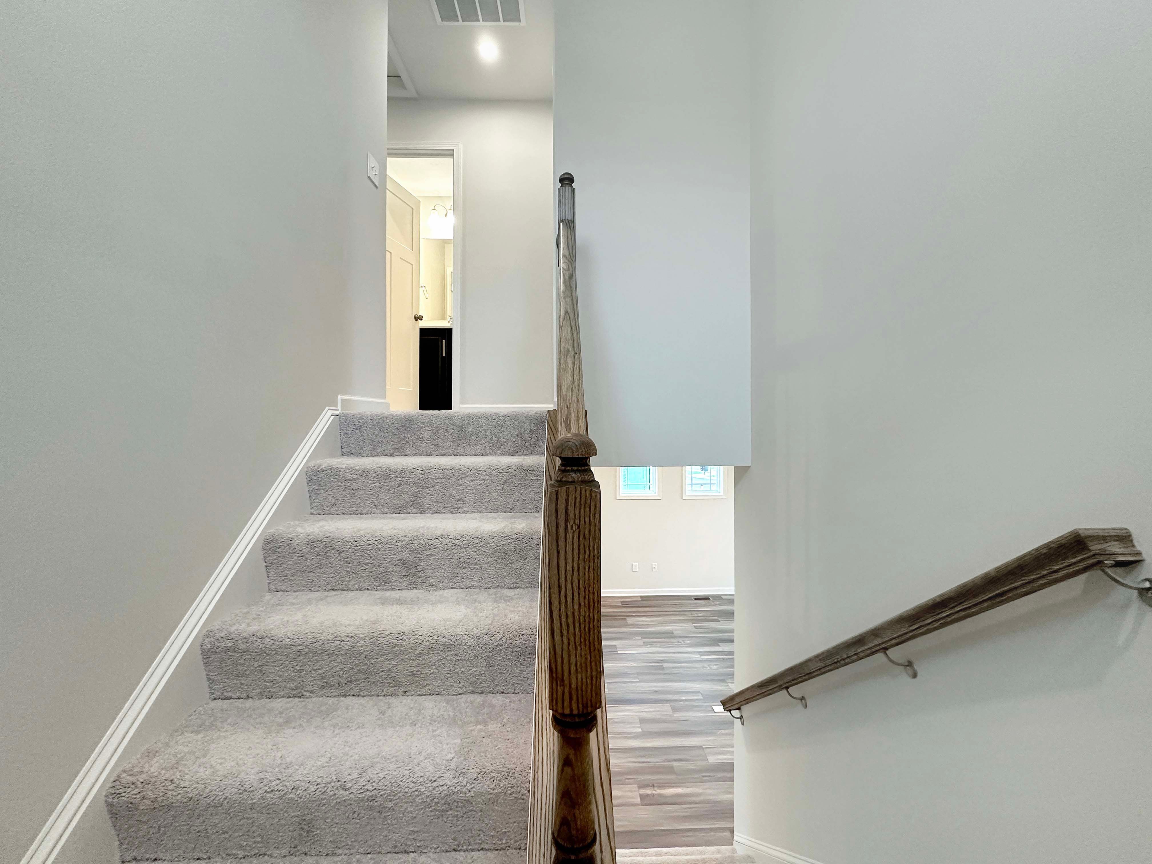 Carpeted staircase with a wooden handrail leading to a hallway with a view into a bathroom and bedroom.