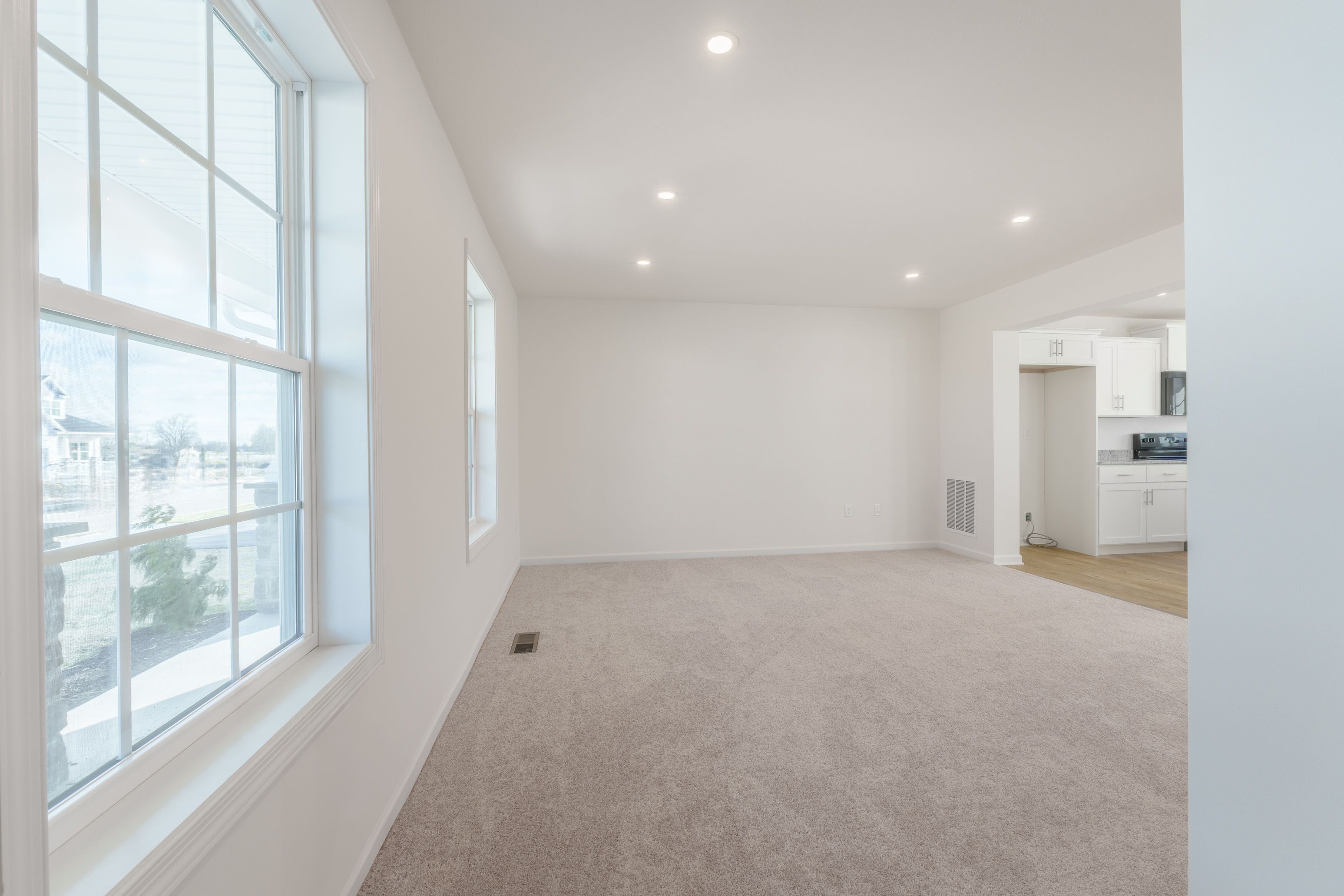 Family room with large windows letting in natural light, light carpet flooring, and neutral walls.