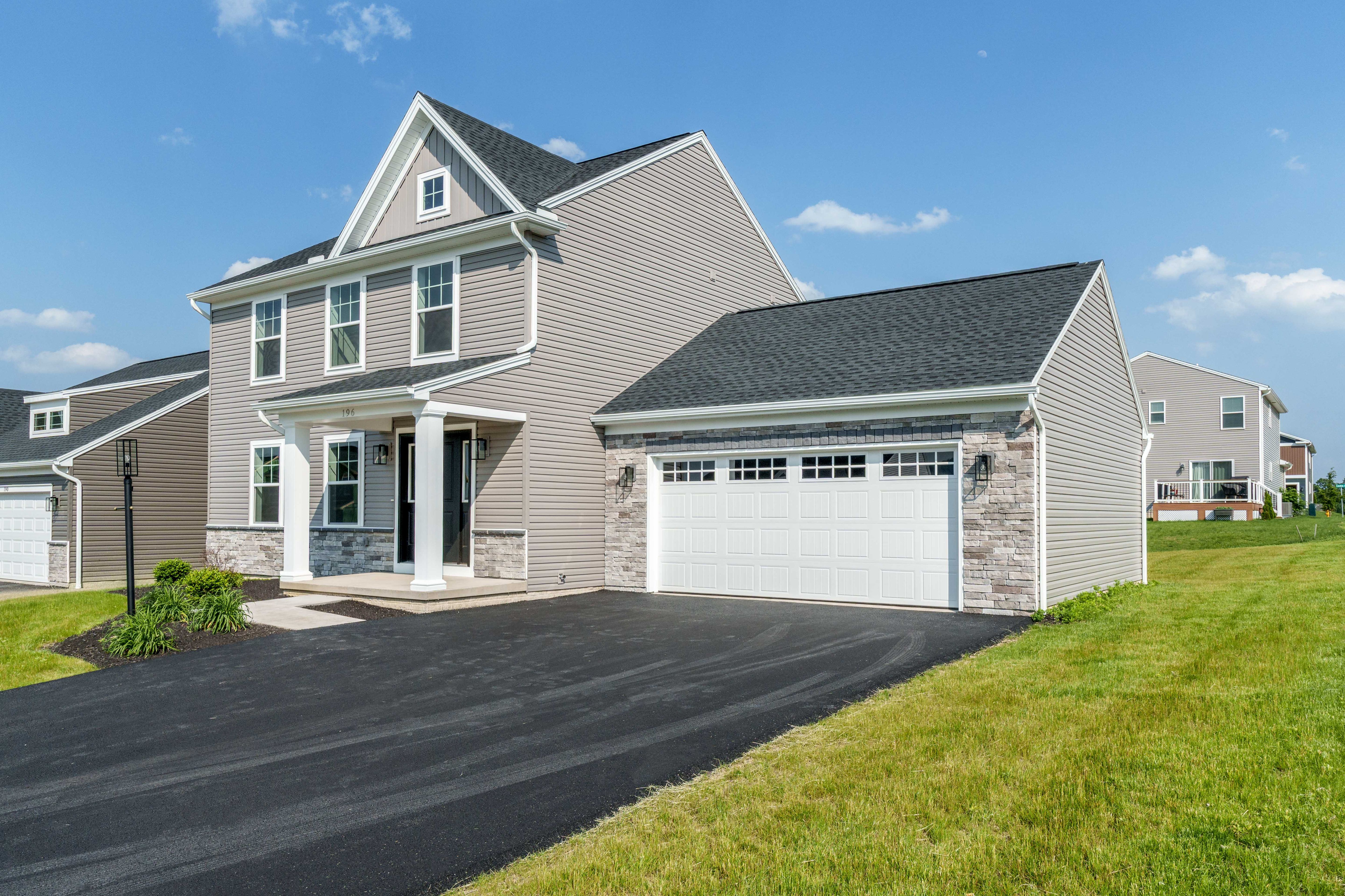 Two-story gray siding home with stone accents, black front door, white trim, and attached two-car garage on a newly paved driveway.