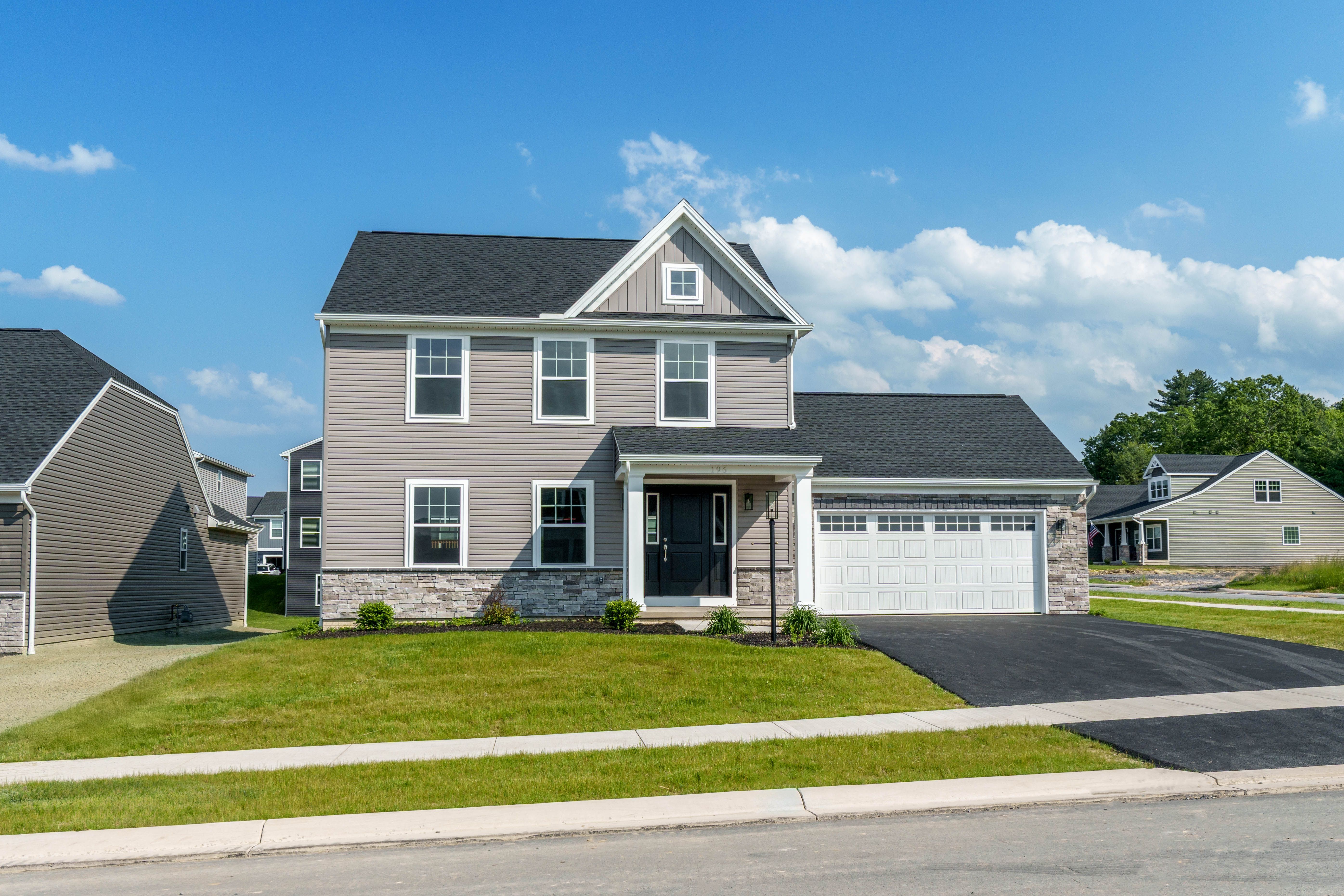 Two-story gray siding home with stone accents, black front door, white trim, and attached two-car garage on a newly paved driveway.