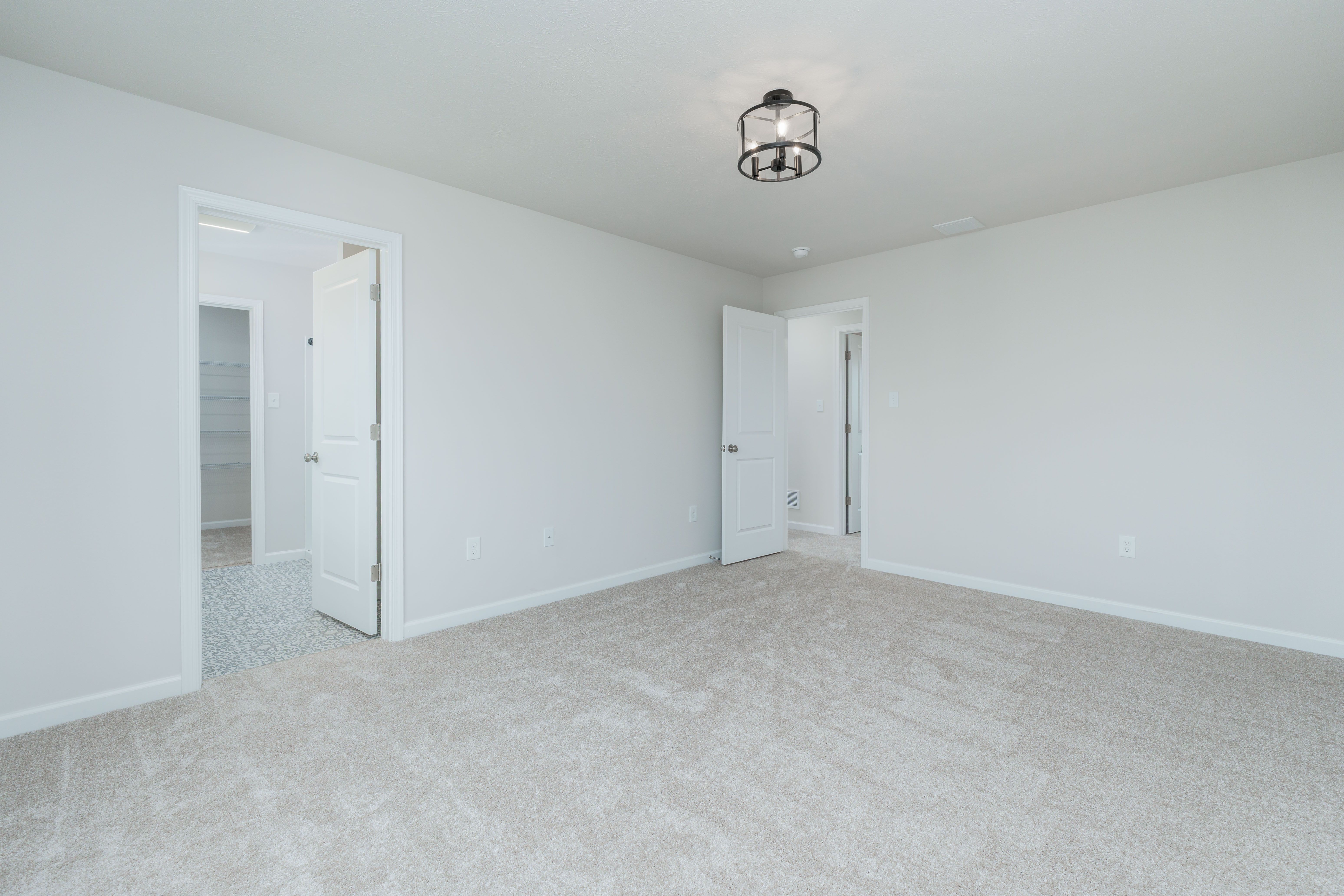 Owners bedroom with carpeted floor, two open doors leading to bath and hallway, and black ceiling light.