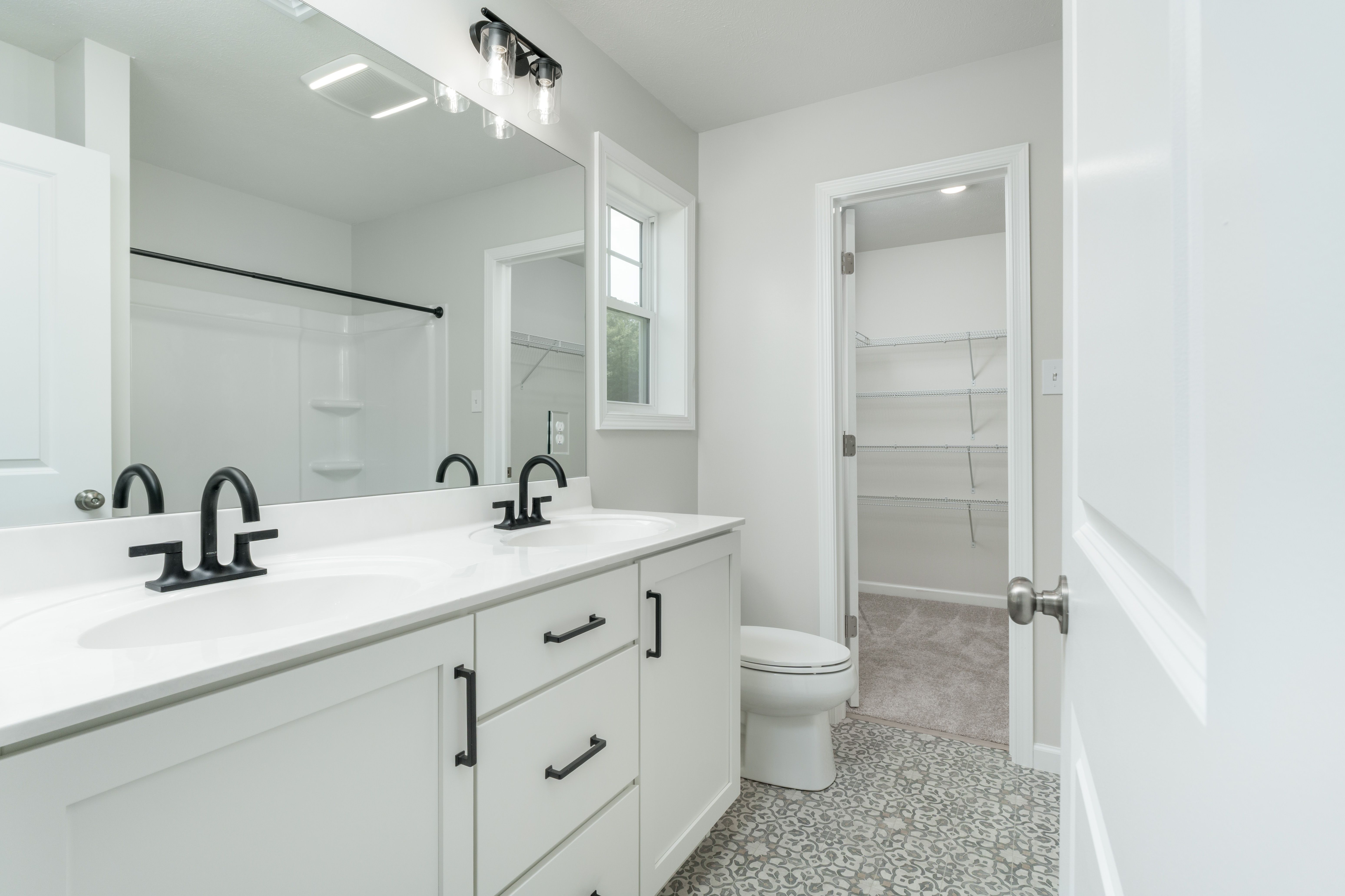 Owners bathroom with dual-sink white vanity, black fixtures, and walk-in closet entrance.