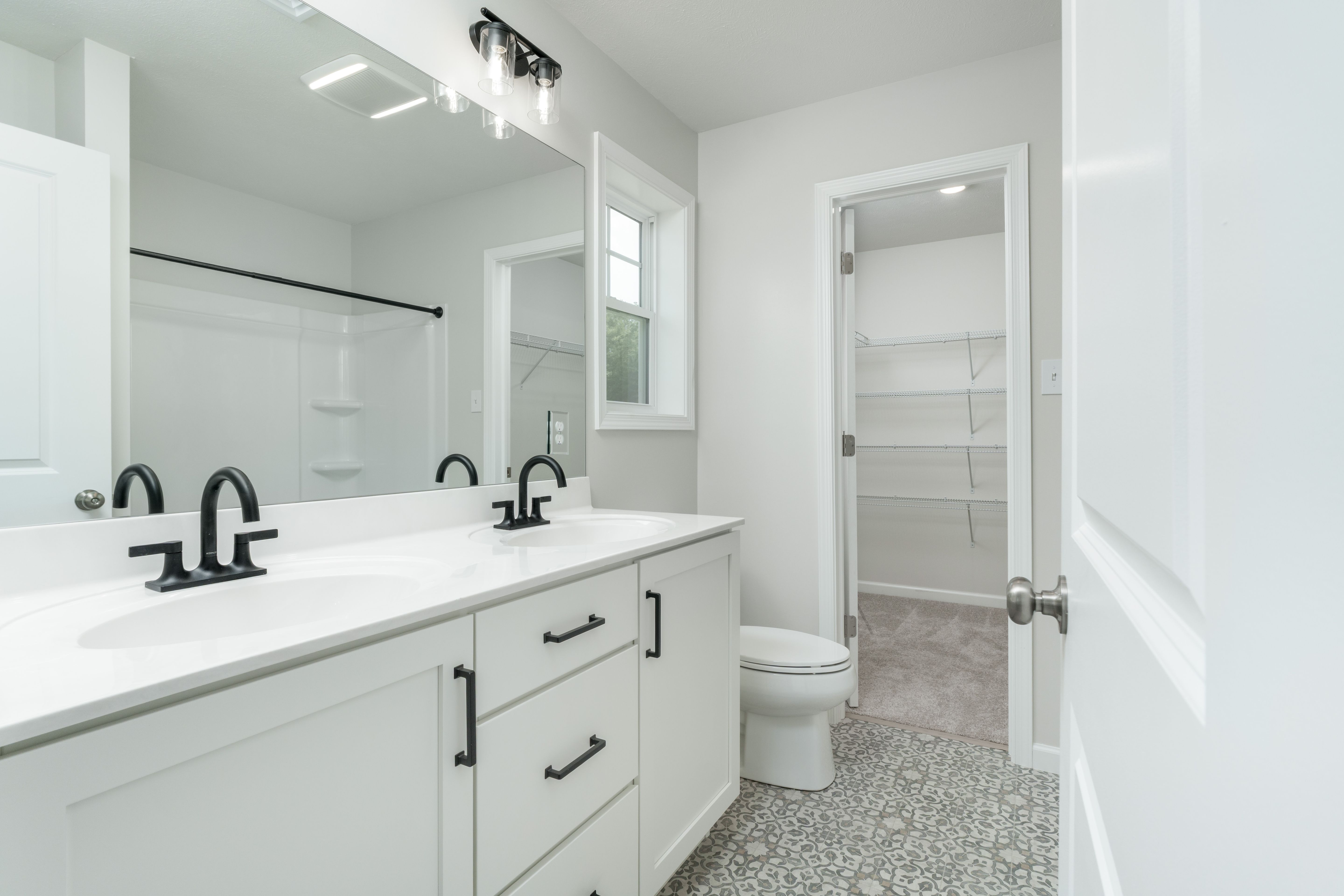 Owners bathroom with dual-sink white vanity, black fixtures, and walk-in closet entrance.