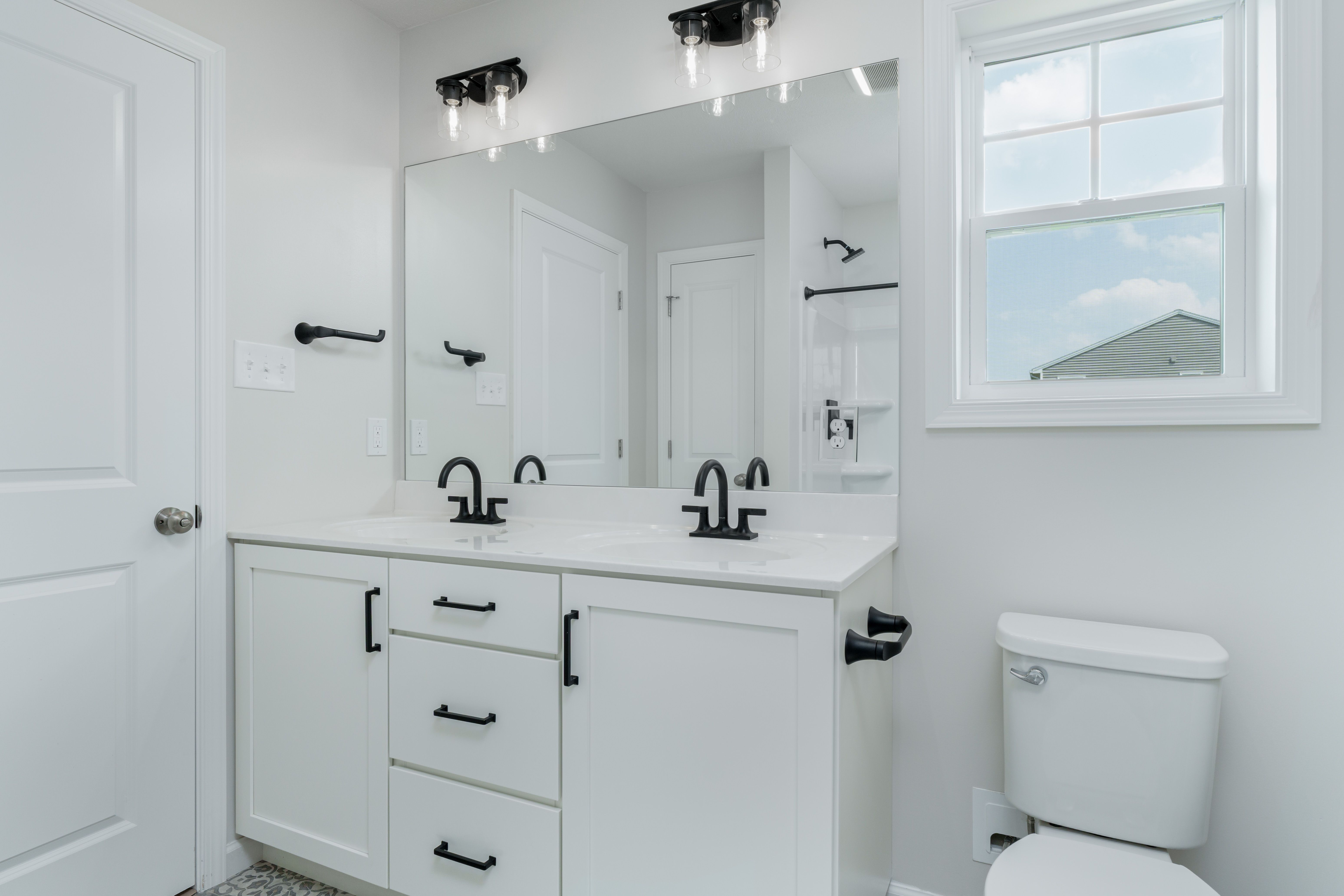 Owners bathroom with large window, dual-sink vanity, and black fixtures beside enclosed shower.