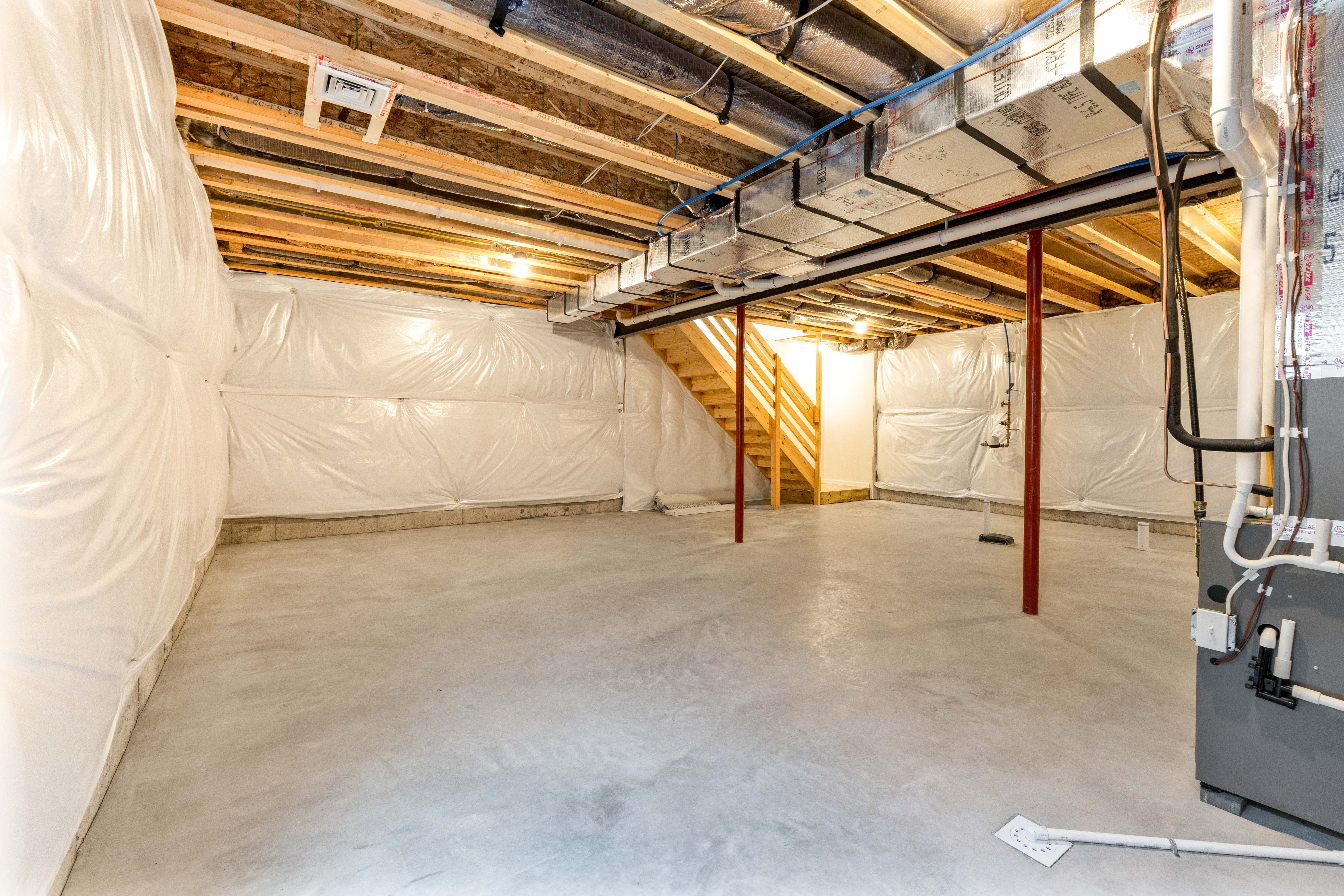 Unfurnished basement corner with exposed ductwork, wood stairs, and white insulated wall wrap along concrete slab.