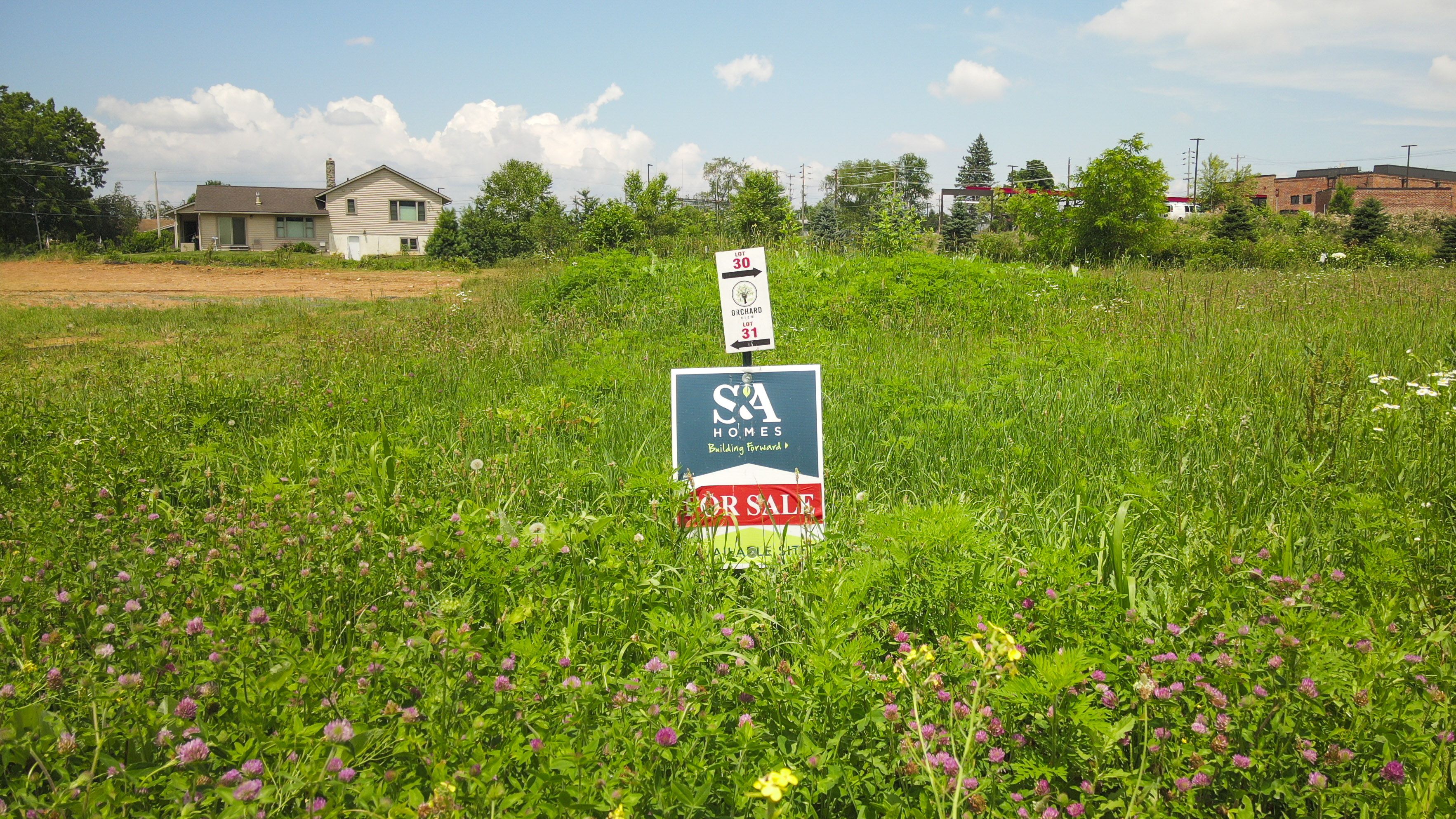 S&A Homes for sale sign on grassy lot with distant home and clear summer sky