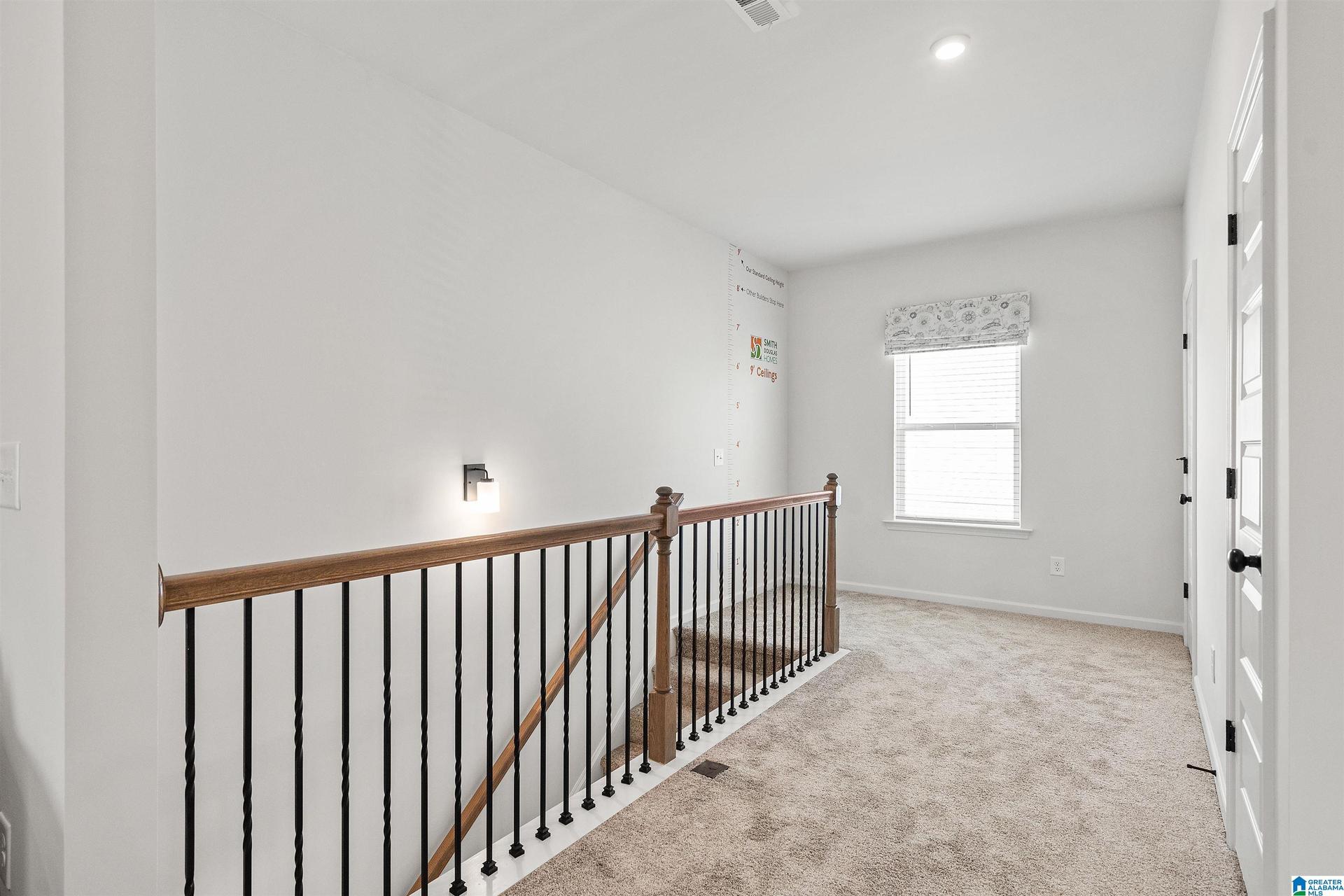 Carpeted hallway featuring a window with blinds and valance, recessed lighting, and a staircase with wood railing and black metal balusters