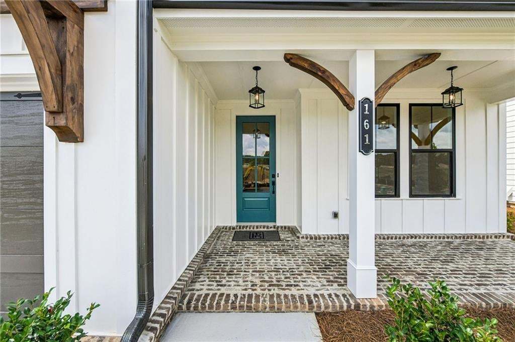 Covered front porch featuring a teal paneled door with glass insert, brick flooring, white board-and-batten siding, and exposed wood beam accents