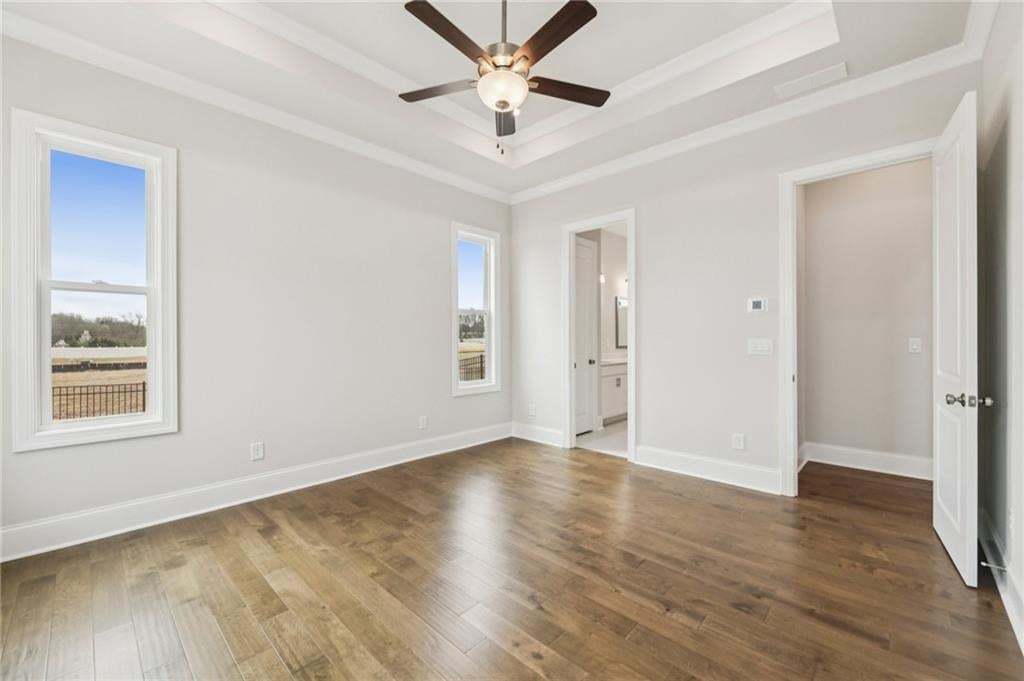 Unfurnished bedroom featuring dark wood-style floors, a ceiling fan, ensuite bath, a tray ceiling, and crown molding