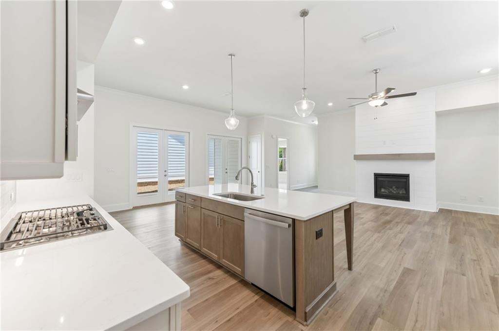 Open-concept living space featuring a central kitchen island with a built-in sink and stainless-steel dishwasher, light wood-finish flooring, a contemporary fireplace with shiplap accent wall and wood mantel, and double French doors to the exterior