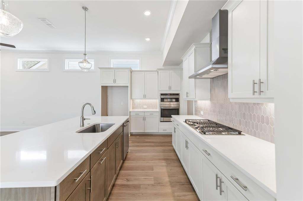 Gourmet kitchen featuring a large central island with an undermount sink, light wood-finish flooring, white shaker-style cabinetry, a built-in oven, and a gas cooktop with a stainless steel range hood