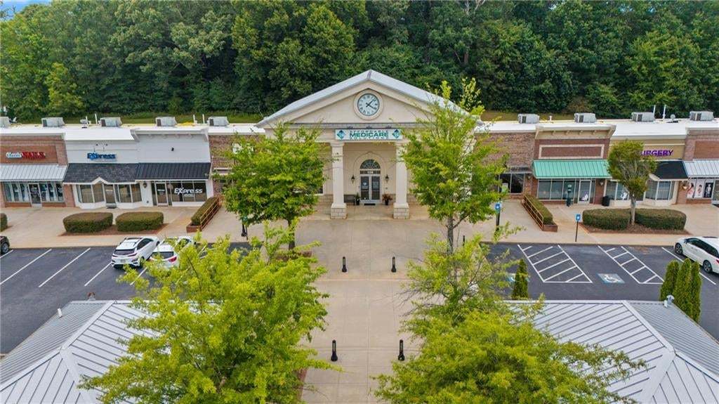 Retail center featuring a central building with a prominent clock, multiple storefronts, paved parking areas, and surrounding mature trees