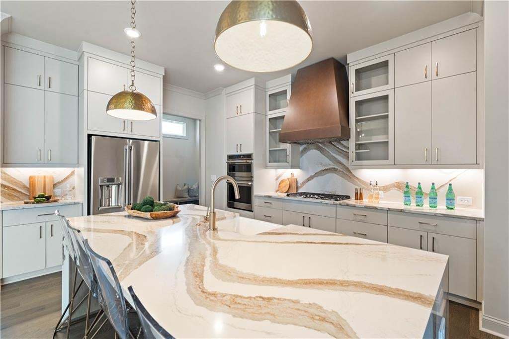 Spacious kitchen featuring a large island with a patterned countertop, light-toned cabinetry, stainless steel appliances, a substantial range hood, and distinctive backsplash detailing
