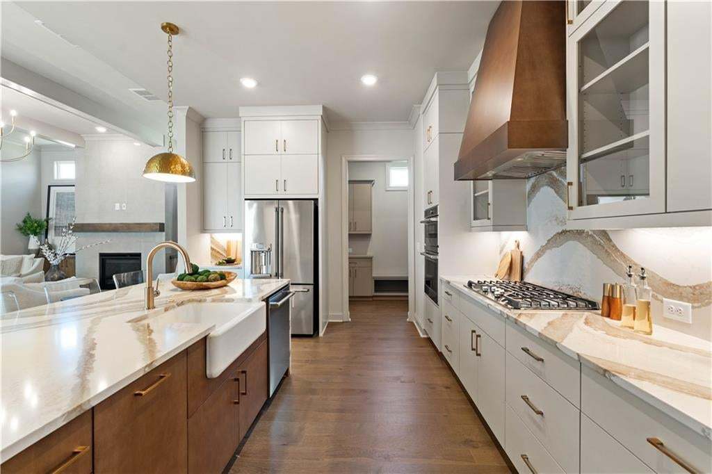 Gourmet kitchen featuring a large island with a farmhouse sink, stainless steel appliances, a gas range with a custom wood-finish range hood, and light-toned countertops with a unique backsplash