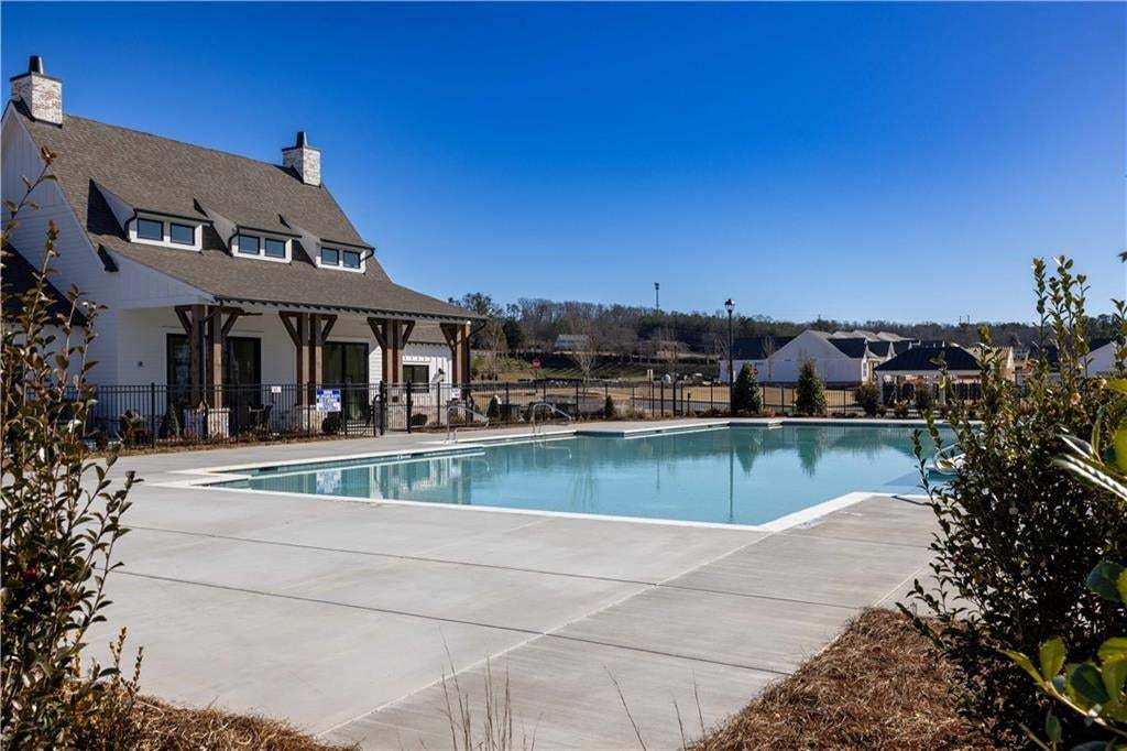 Community pool with concrete decking, surrounded by a black metal fence, adjacent to a clubhouse featuring a covered porch with exposed wood beams and white siding