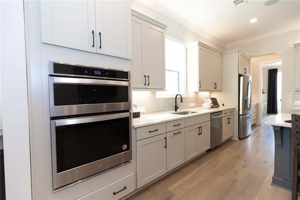 Stainless steel double ovens, wood-finish flooring, light-toned cabinetry with dark hardware, recessed lighting, and a subway tile backsplash