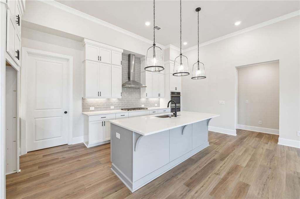 Kitchen featuring a large island with sink, white shaker-style cabinetry, stainless steel range hood, light wood-finish flooring, and pendant lighting