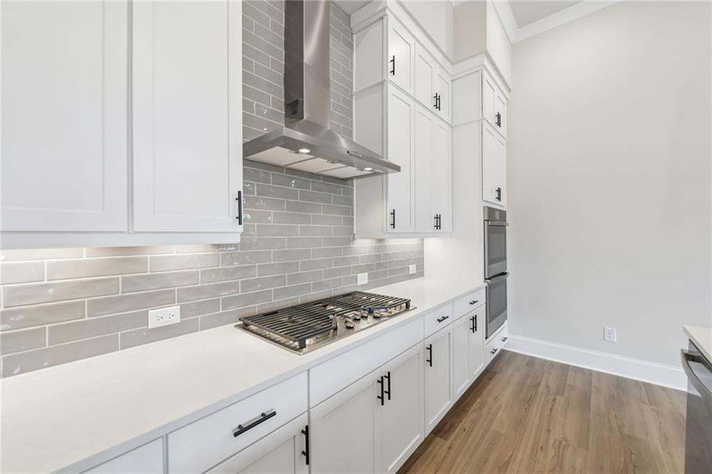 White cabinetry, light-toned subway tile backsplash, stainless steel range hood, built-in double oven, and wood-finish flooring